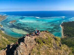 A group of customers on the summit of le morne brabant, hands in the air, celebrating their achievement. The scenery around them is breathtaking, pristine nature, crystal clear lagoon that fades with the clear blue sky, on the left the arid vegetation of Baie du Cap, and on the right the filaos of Le Morne.