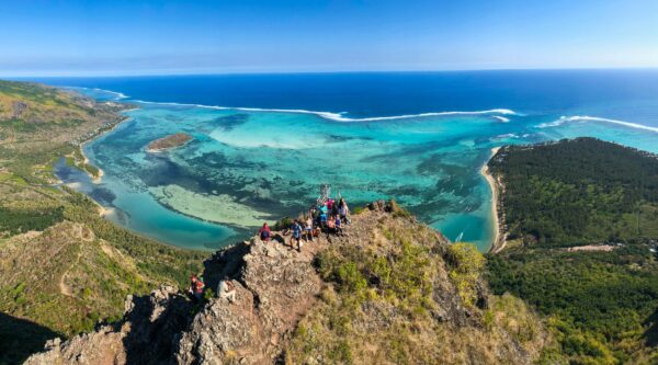 A group of customers on the summit of le morne brabant, hands in the air, celebrating their achievement. The scenery around them is breathtaking, pristine nature, crystal clear lagoon that fades with the clear blue sky, on the left the arid vegetation of Baie du Cap, and on the right the filaos of Le Morne.